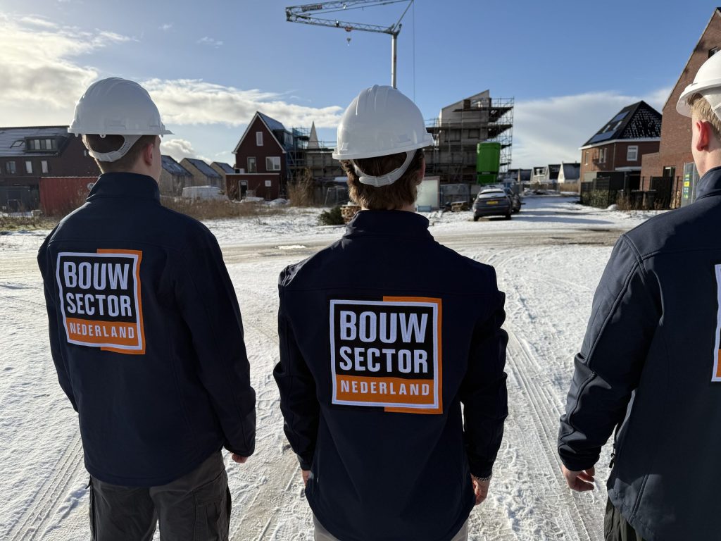 Drie vakmensen in bedrijfsjas kijken samen naar een woning met steigers op een winterse bouwplaats